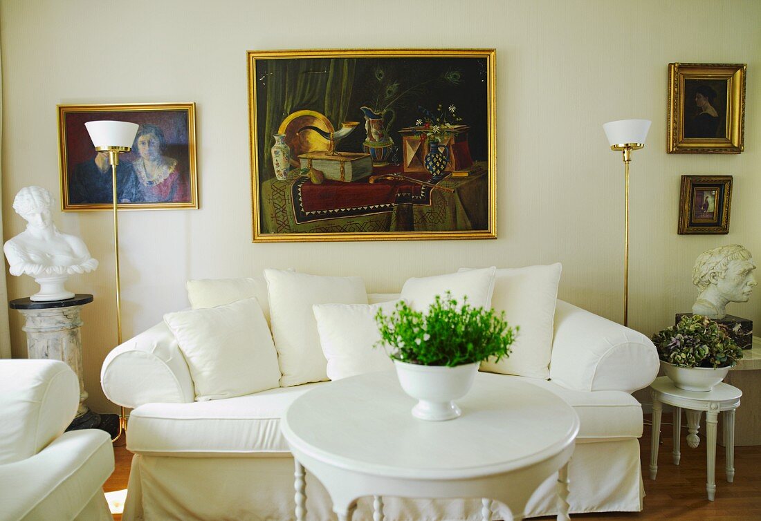 White sofa with valance and vintage coffee table between antique busts and against wall with paintings from various periods