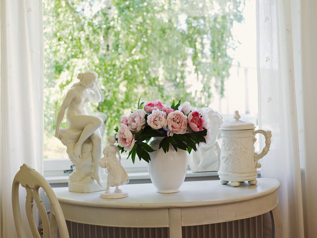 Bouquet of peonies between figurines and beer stein on white, semi-circular console table below window