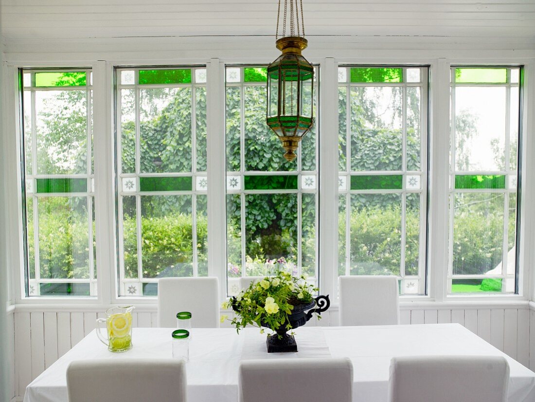 Table with white cloth and white chairs in dining room with Art Deco window