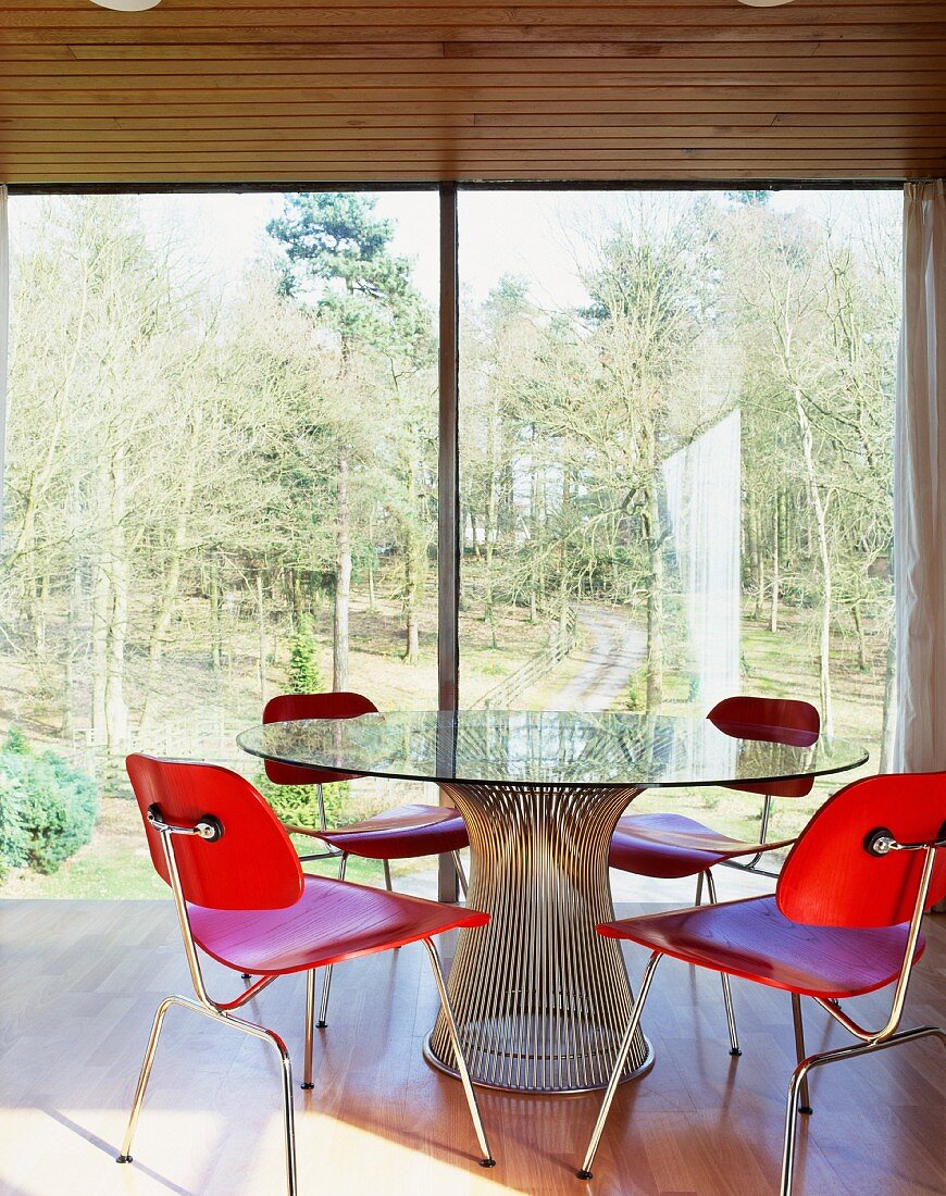Rot gebeizte Holzstühle um Glastisch vor raumhohen Terrassenfenstern mit Ausblick