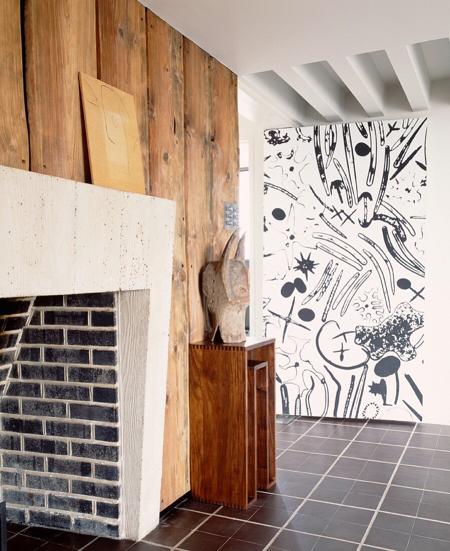 Brick fireplace on wood-clad wall opposite wall with black and white patterned wallpaper