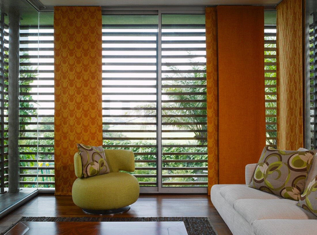Kleiner, grüner Polstersessel vor Sonnenschutzlamellen mit Kissenmuster und orangefarbenen Schiebevorhängen im 70er Stil