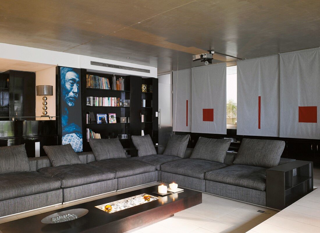 Graue Sofalandschaft mit einzelnen Rückenkissen und Designer Couchtisch in Wohnzimmer mit Beamer unter Betondecke und Schiebegardinen