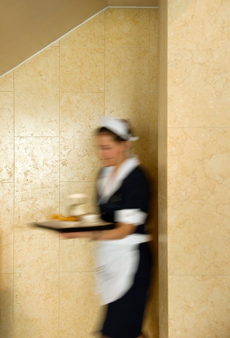 Blurred waitress in traditional uniform hurrying with full tray against marble-clad, sand-coloured wall