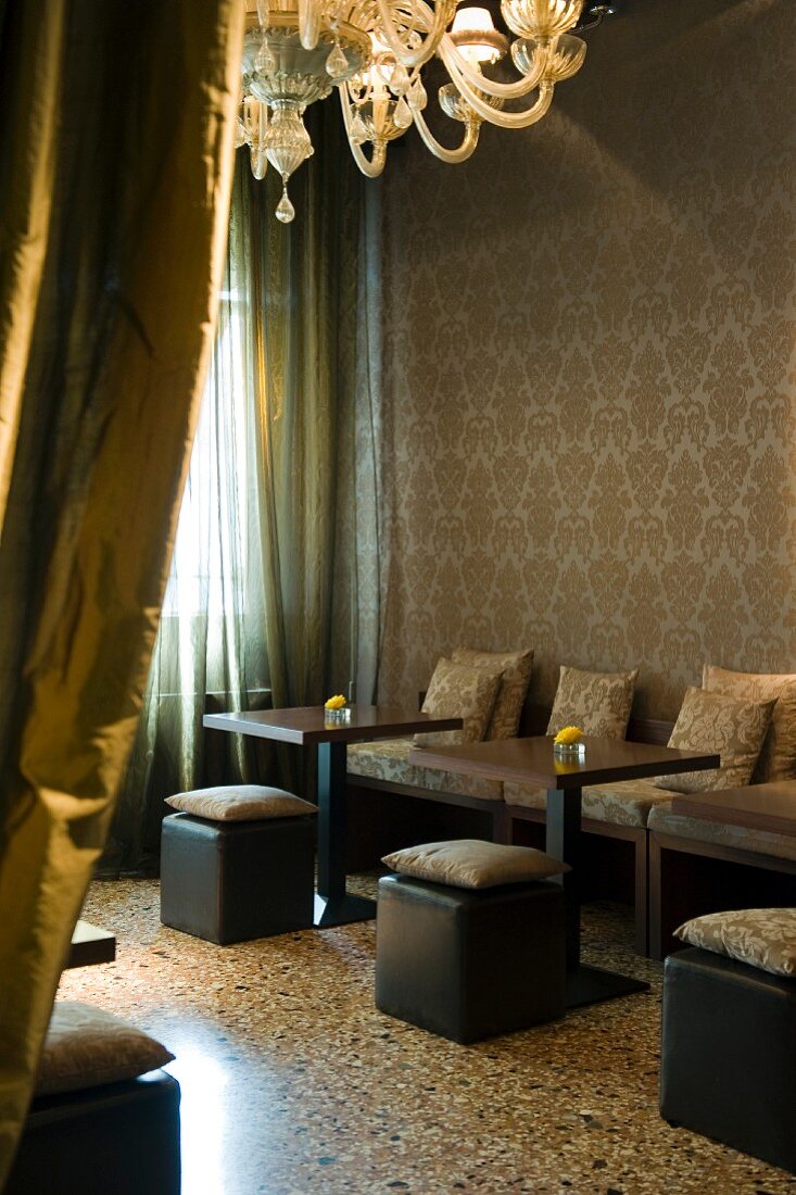 View past mustard-coloured silk curtain into vintage Venetian-style hotel lobby with cushions on black cubic stools and terrazzo flooring