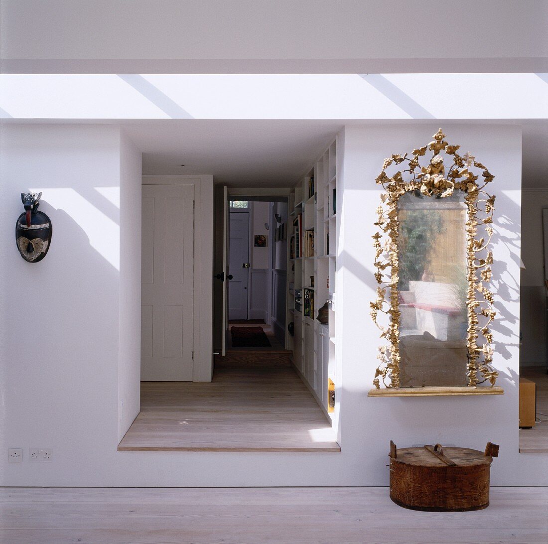 Blick in erhöhten Flur mit offener Wohnungstür zwischen exotischer Maske und Spiegel mit goldenem Rahmen in Form von Rankblättern