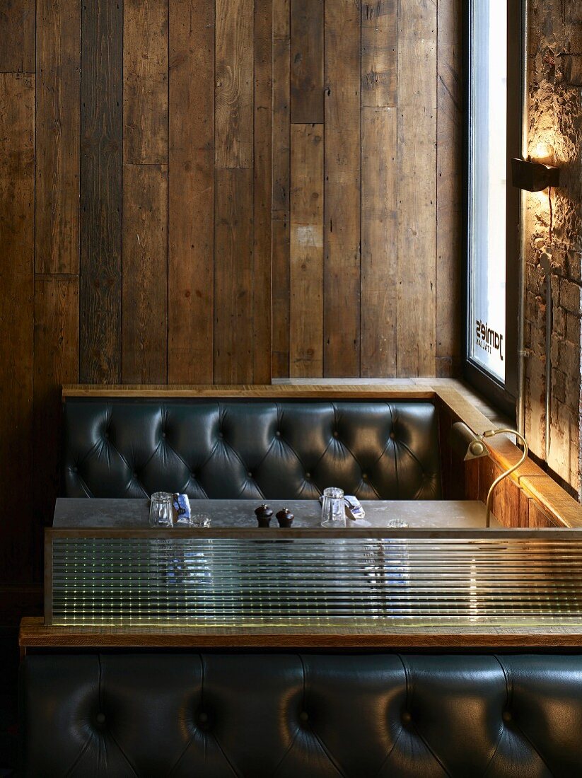 Lounge-Nischen im Vintage Look - schwarze Lederpolsterbänke und messingfarbene Retro-Tischlampe vor alter Holzwand