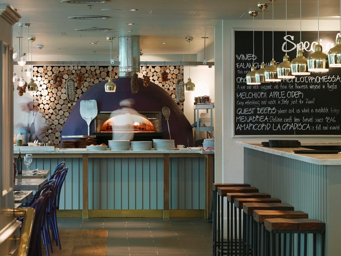 Restaurant in London with modern pizza oven in front of wood stack and bar with pendant lights in front of specials board