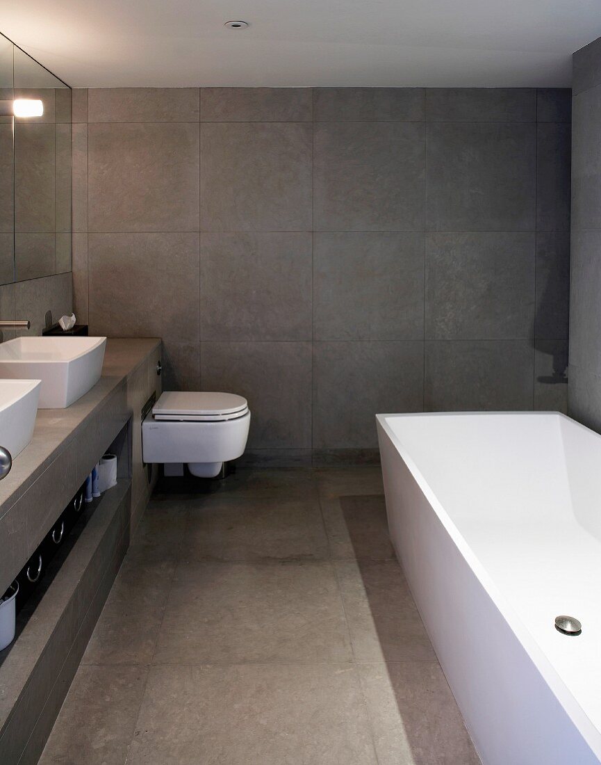 Bathroom tiled in grey with bathtub, washstands & toilet
