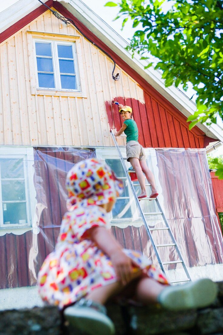 Junge Frau streicht die Fassade des Hauses, kleine Tochter sieht zu