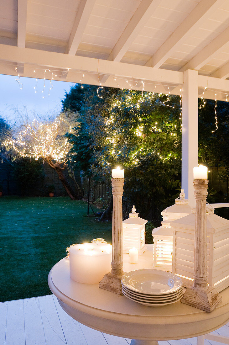Festliche Stimmung auf Veranda mit beleuchteten Laternen neben Kerzenständern auf Tisch und Blick auf Lichterketten in den Bäumen
