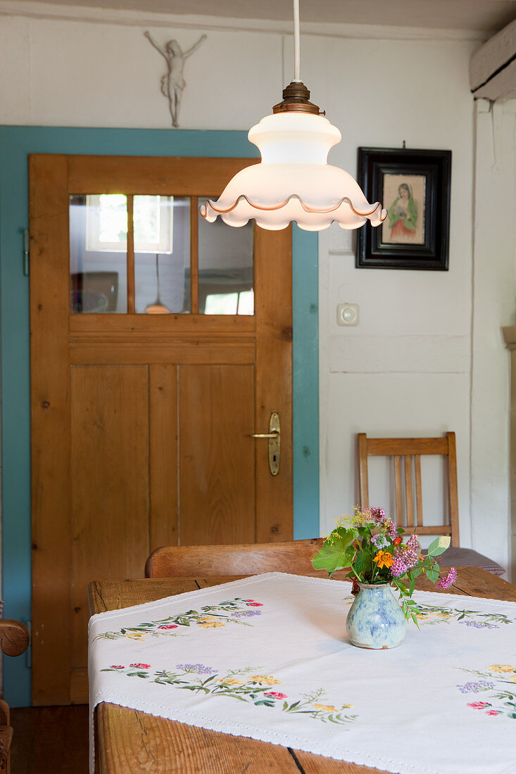 Embroidered tablecloth and vase of flowers on kitchen table below vintage pendant lamp opposite interior door with glass panels