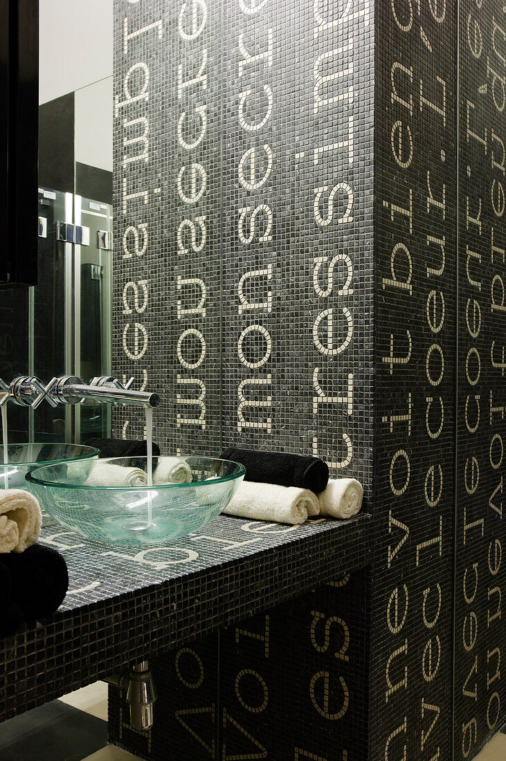 Fliessendes Wasser aus Wandarmatur in Glasschüssel auf einem Designer Waschtisch mit Fliesenmosaik aus dunklem Hintergrund mit weissen Schriftzügen