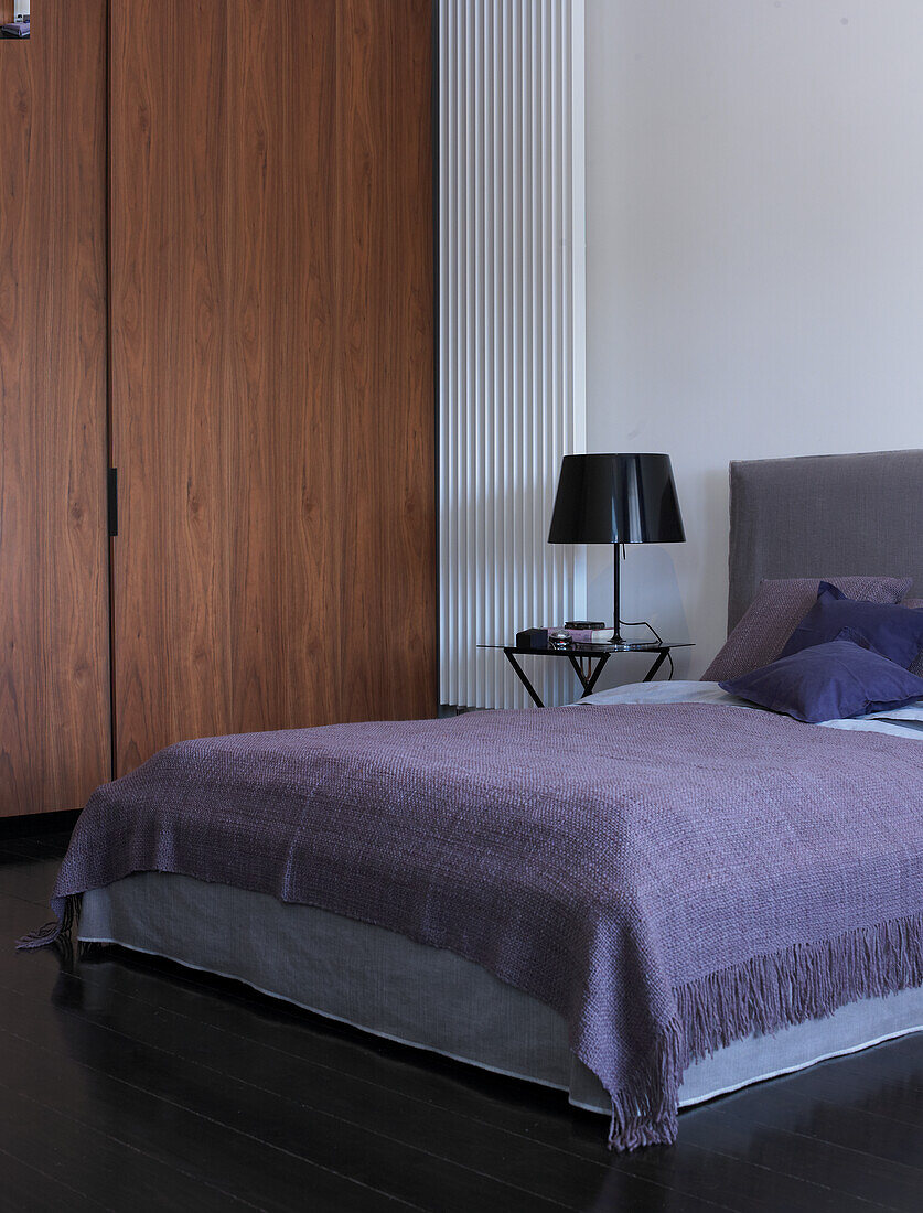 Bed with purple blanket and black table lamp next to wooden wardrobe