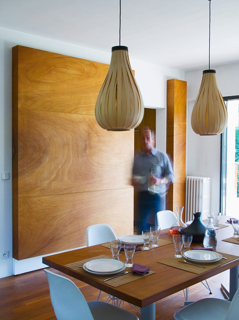 Mann deckt Tisch in modernem Esszimmer mit bauchigen Hängelampen aus Holzstreifen
