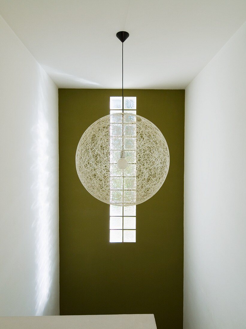 Kugelförmige Hängelampe aus hellem Geflecht im Designerstil vor grüner Wand mit Glasbausteinstreifen im Treppenhaus