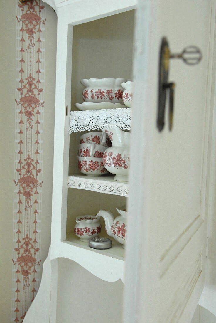 Weisses Küchenbuffet mit offener Tür und Blick auf traditionelles Service mit rot-weißem Blumenmuster