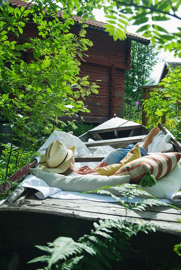 Frau liegt gemütlich auf Kissen und Polstern in ausrangiertem Holzboot im Garten neben schlichtem Holzhaus
