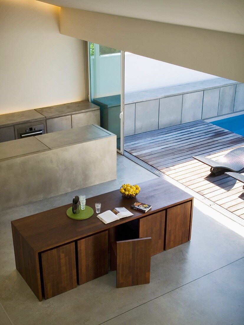 View from above of cubist, designer dining table and chairs and monolithic kitchen island in front of open sliding door leading to terrace with wooden deck and pool