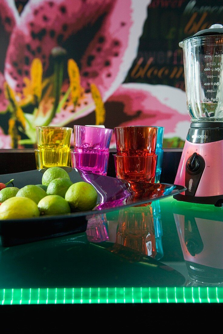 A bowl of limes next to coloured glasses and a retro blender on the illuminated frosted glass counter of a breakfast bar
