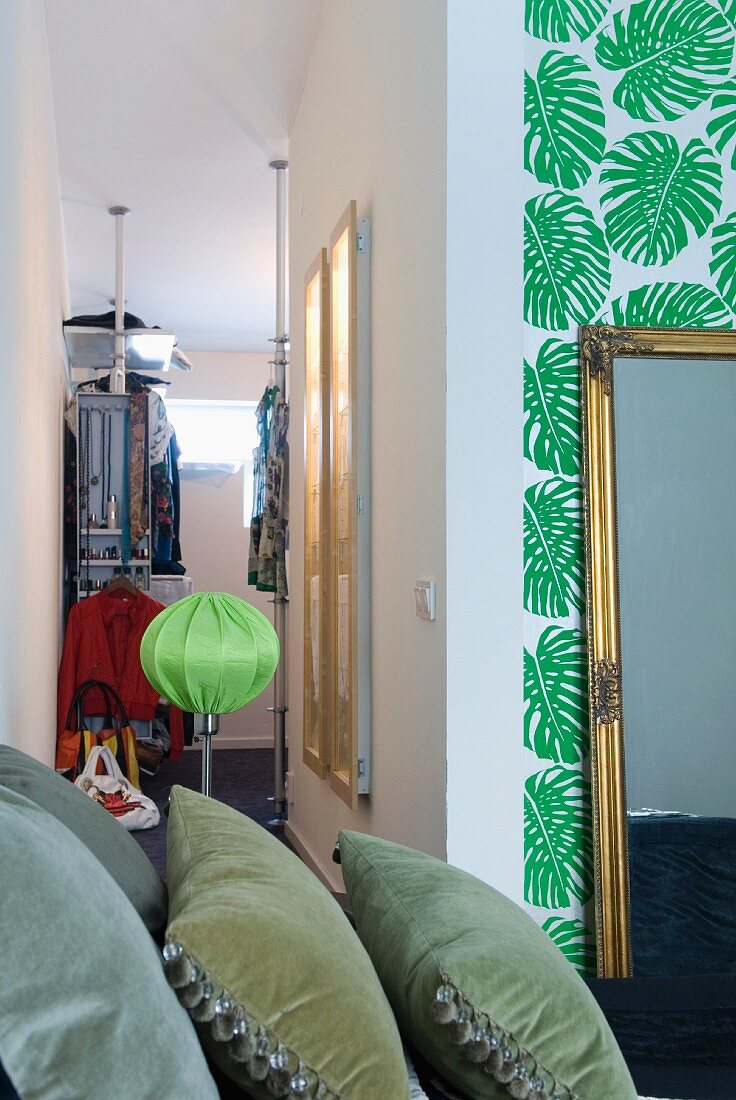 Blick über Samtkissen hinweg in den begehbaren Kleiderschrank eines stilgemixten Schlafzimmers mit Palmblatttapete