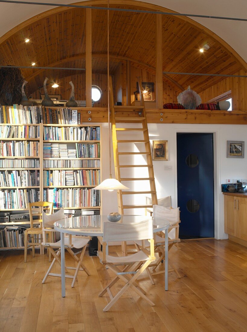 Dining area with bookcase next to ladder leading to comfortable sleeping gallery with wood-panelled arched ceiling