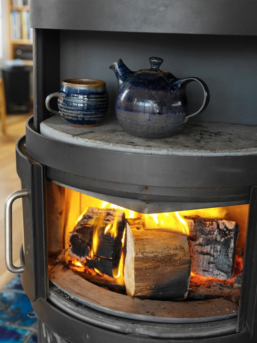 Blue-glazed ceramic teacup and teapot on warming plate of wood-burning stove with roaring fire