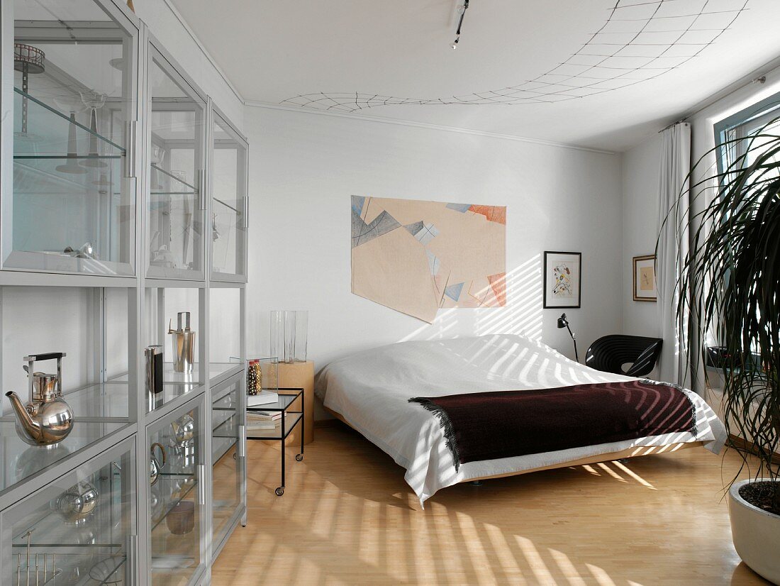 Schlafzimmer mit einfachem Doppelbett und moderner Glasvitrine mit SAmmlung verschiedener Gefäße