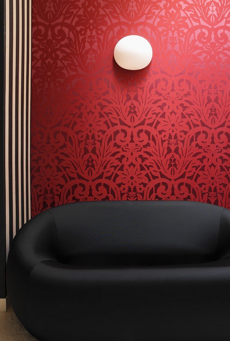Black designer sofa in front of red wallpaper with traditional, floral pattern