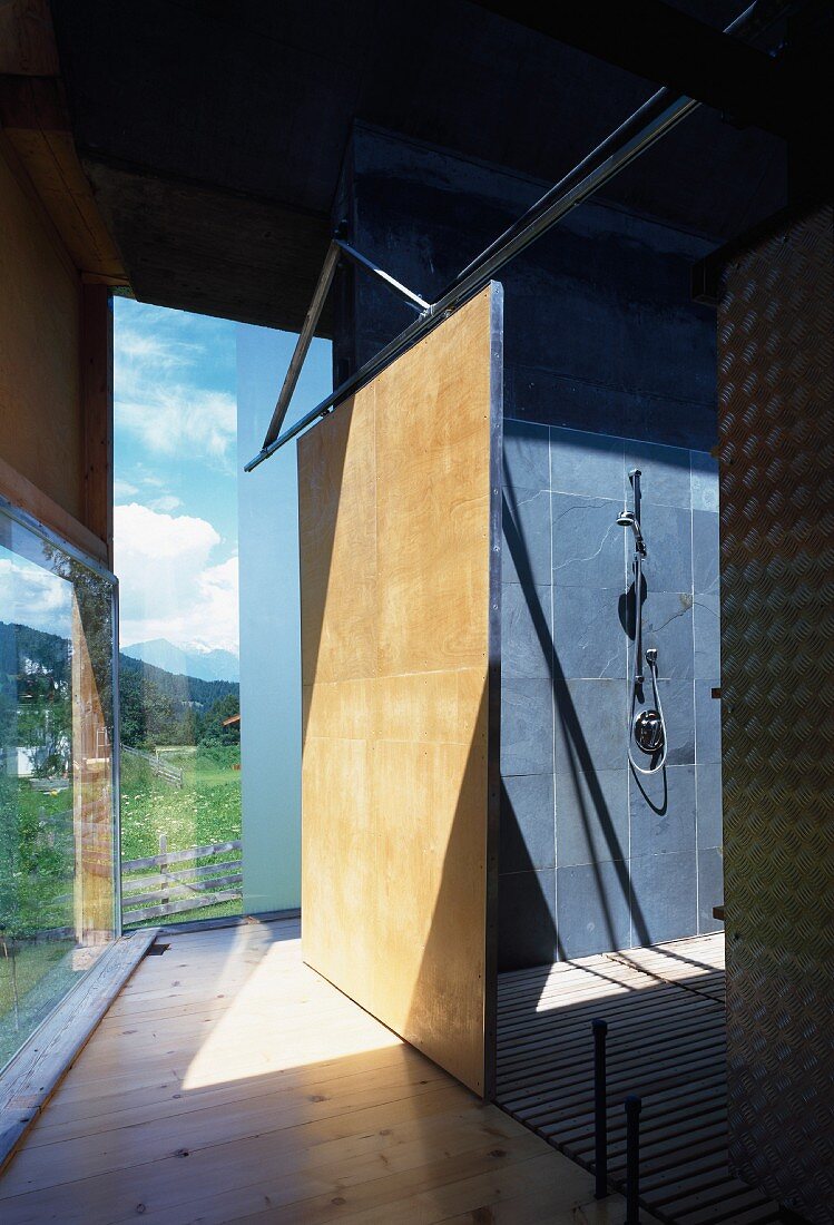 Gang an Glasfassade mit Blick durch Schiebetür in Duschraum mit graublauen Wandfliesen und Holzrost