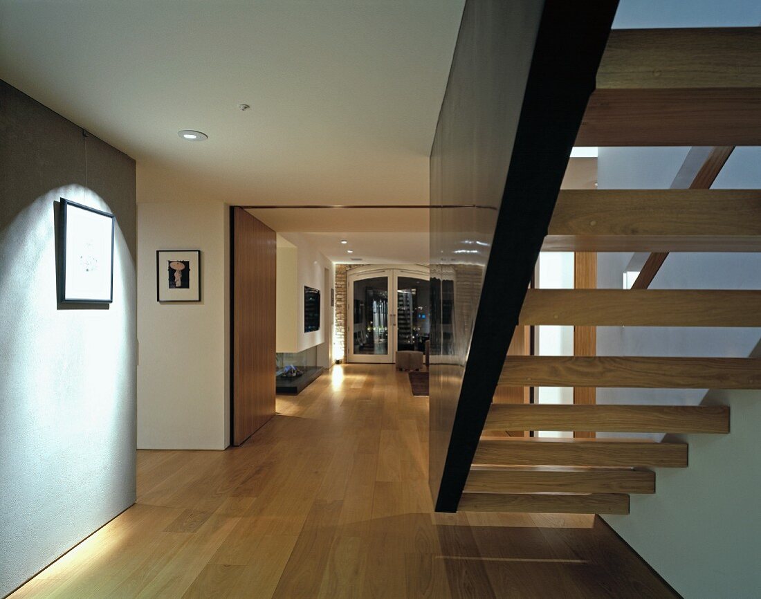 Floating staircase between wall and dark stringer element in wide hallway with view of open-plan living space
