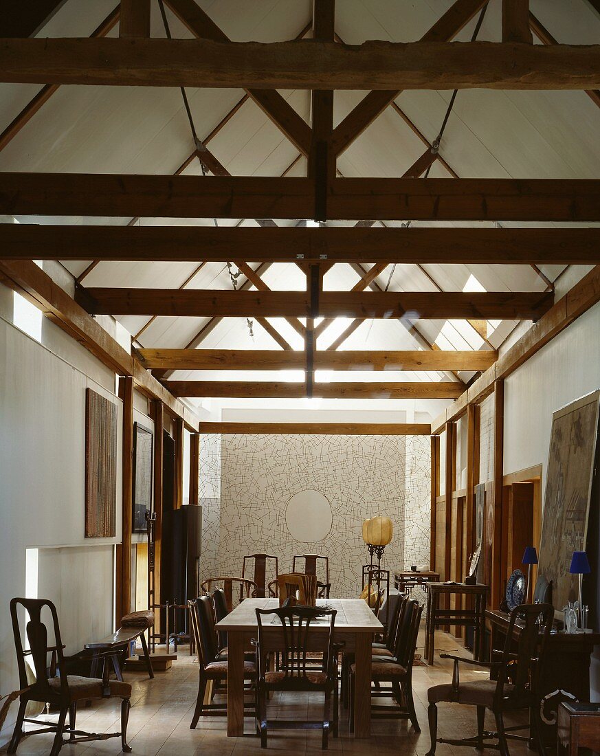 Lange Esszimmertafel mit zahlreichen antiken Holzstühlen und flächiger, filigraner Drahtskulptur unter moderner Dachstuhlkonstruktion