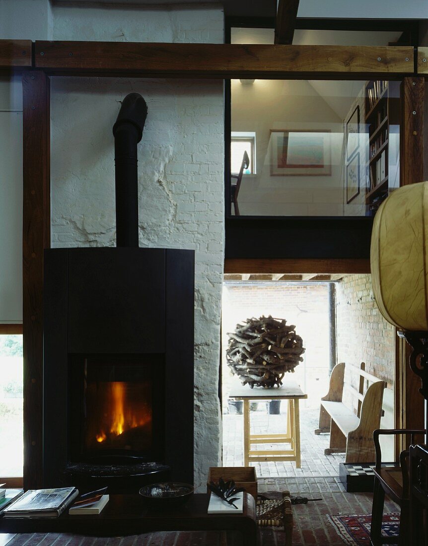 Lit wood-burning stove in double-height living space with view of spherical wooden sculpture and library level with glass wall above