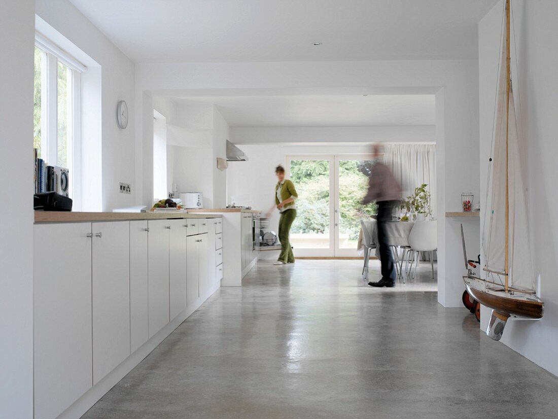 Woman in front of long kitchen unit and man in dining area of modern room with concrete floor