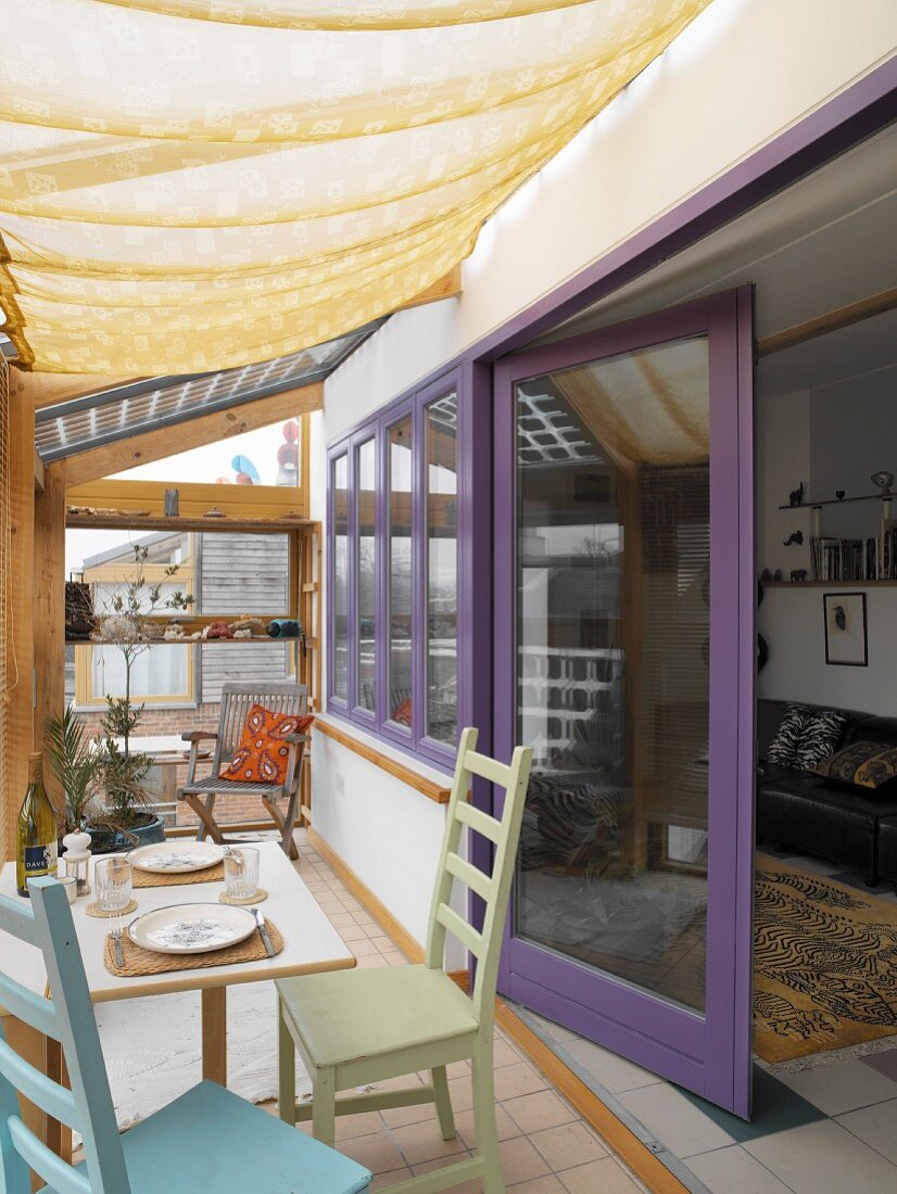 Conservatory with fabric sunshade and simple wooden furniture in front of living room with lilac window frames