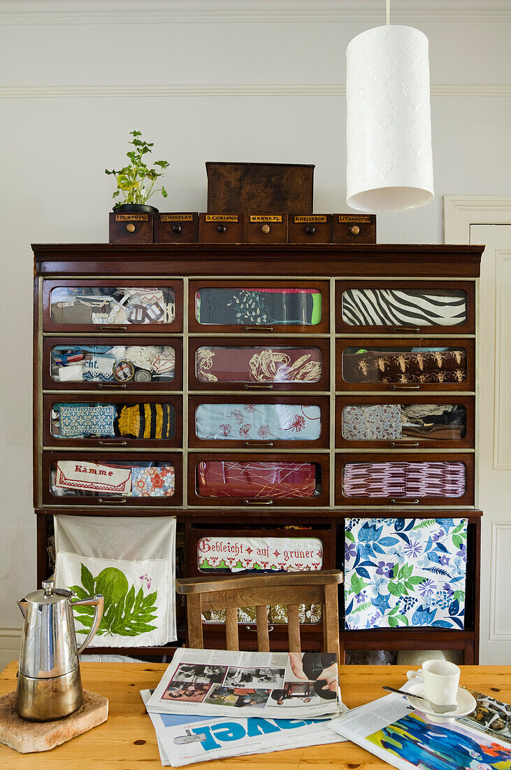 Old wooden cupboard with patterned fabrics, wooden table with coffee pot, coffee cup and newspapers