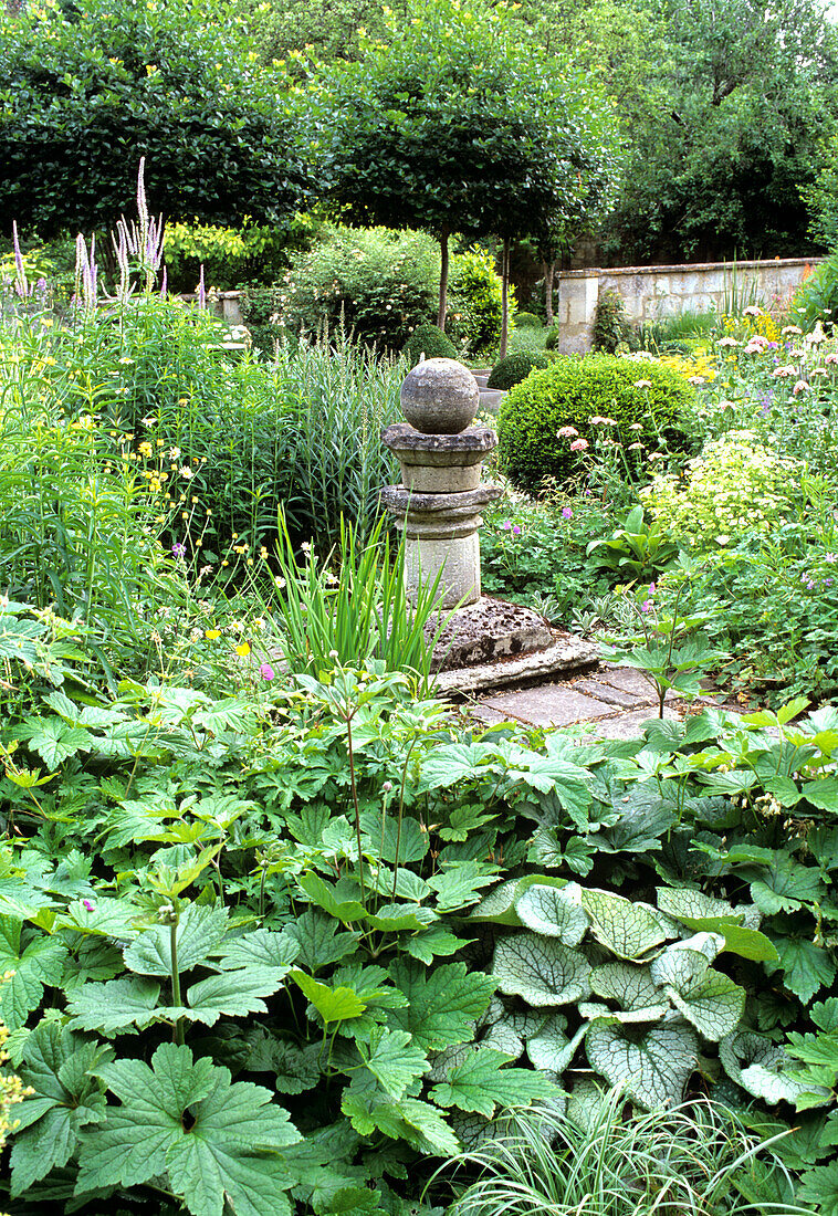 Steinsäule umgeben von vielfältigen Grün- und Blühpflanzen im Garten