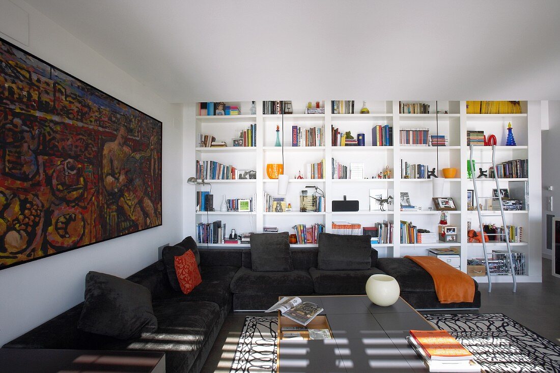 Modernes Wohnzimmer mit schwarzer Eckcouch; dahinter eine breite, weiße Bücherwand und ein großes Gemälde