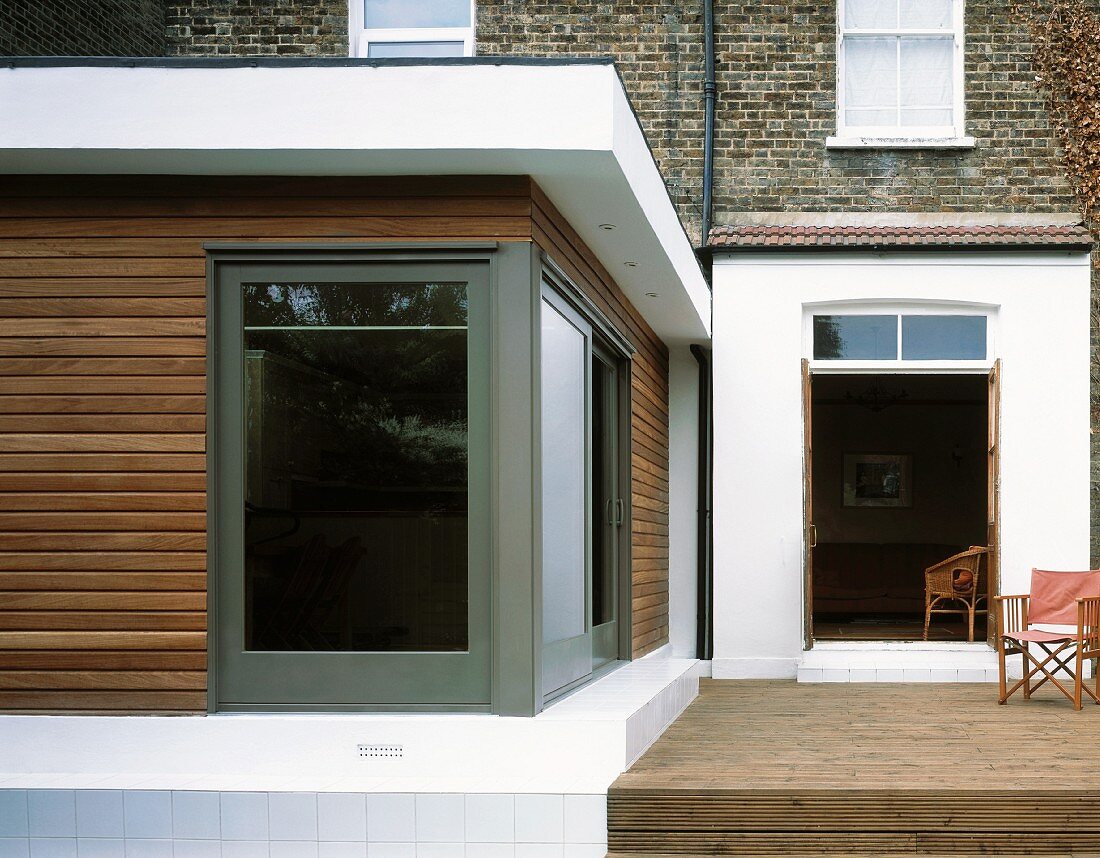 Erdgeschossiger moderner Anbau mit Holzfassade und raumhohem Fenster neben Terrasse mit Holzboden