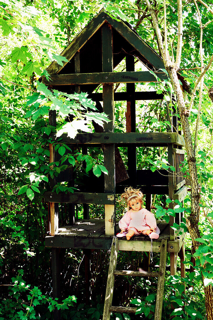 Doll on wooden ladder of a tree house surrounded by green leaves