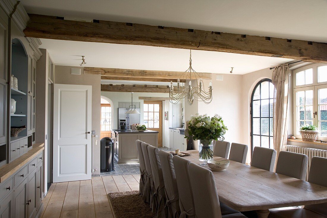 Long table and loose-covered chairs in country-house dining room with adjoining, open-plan kitchen