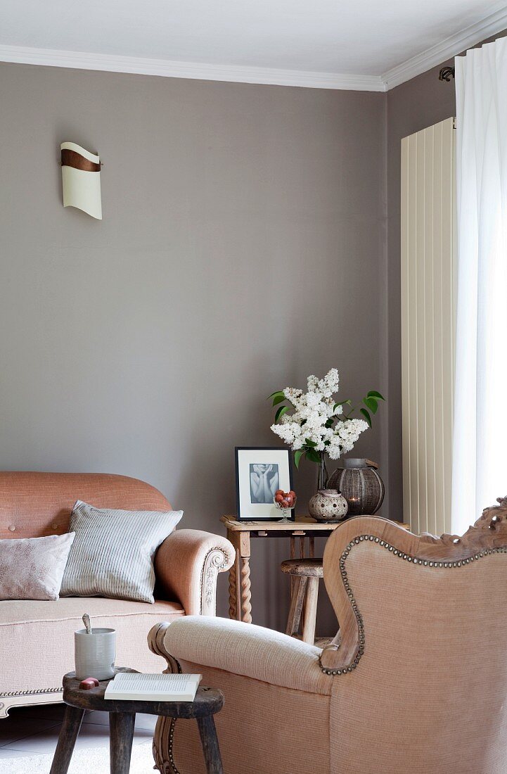 Rustic wooden stool next to dusky rose Rococo armchair and matching sofa against wall painted an elegant grey