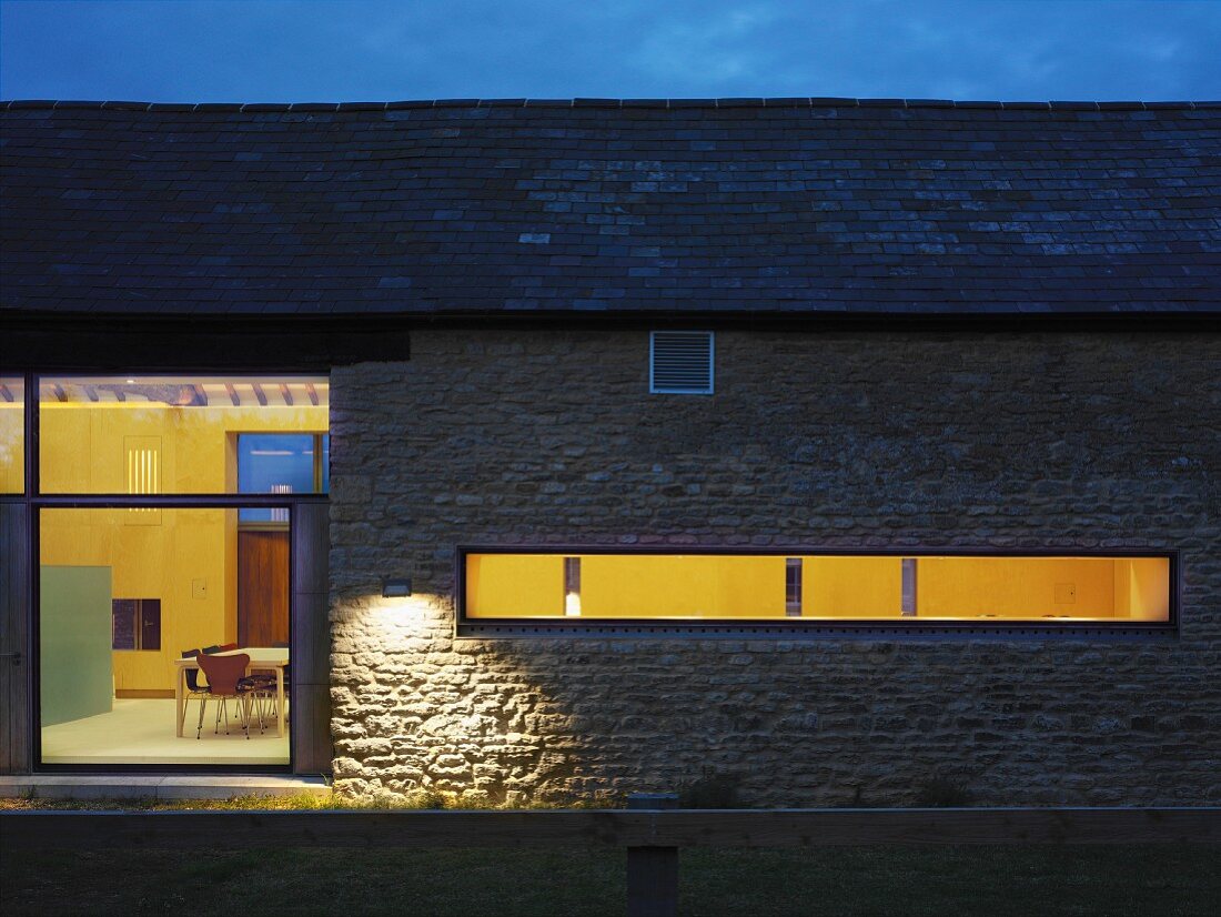 Renoviertes Bauernhaus mit Steinmauer in Abendstimmung und Blick durch Fenster in beleuchteten Innenraum