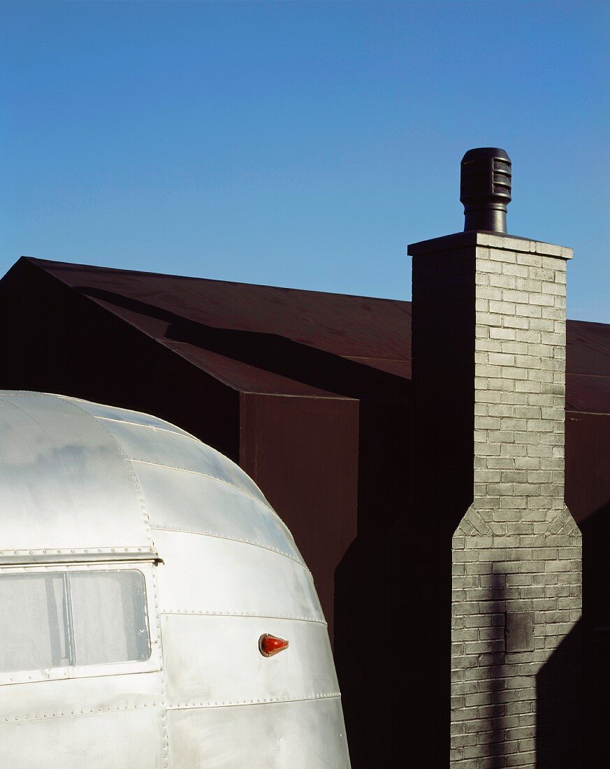 Metal-clad, retro caravan in front of house with black outer shell and chimney