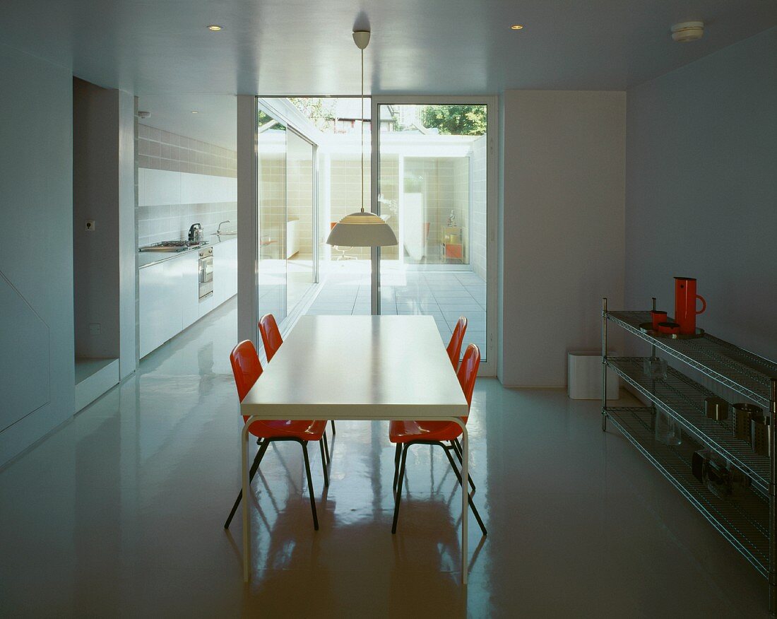 White dining table and orange shell chairs in minimalist room with view of courtyard