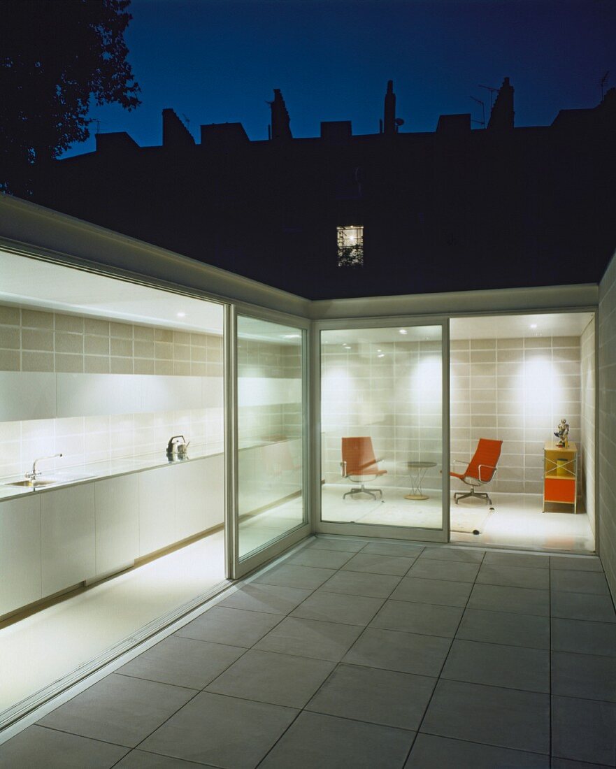 Innenhof mit weissen Bodenfliesen und Blick durch offene Terrassenschiebetüren in beleuchtete Küche und Chillecke