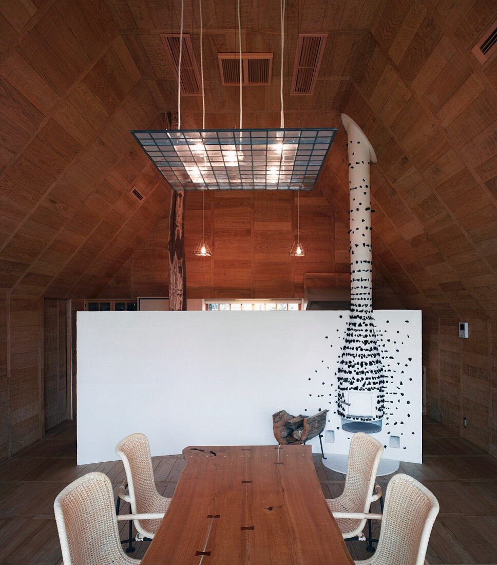 Esstisch aus Holz mit Rattanstühlen im holzverkleideten Wohnraum und Blick auf weissen, gebogenen Raumteiler