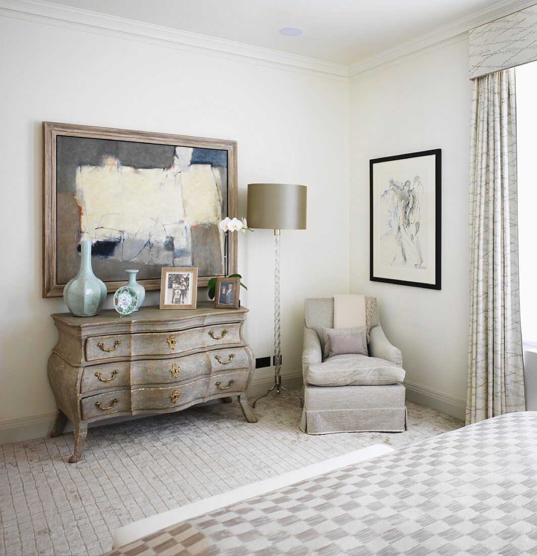 Light bedroom with Rococo chest of drawers next to light grey upholstered armchair