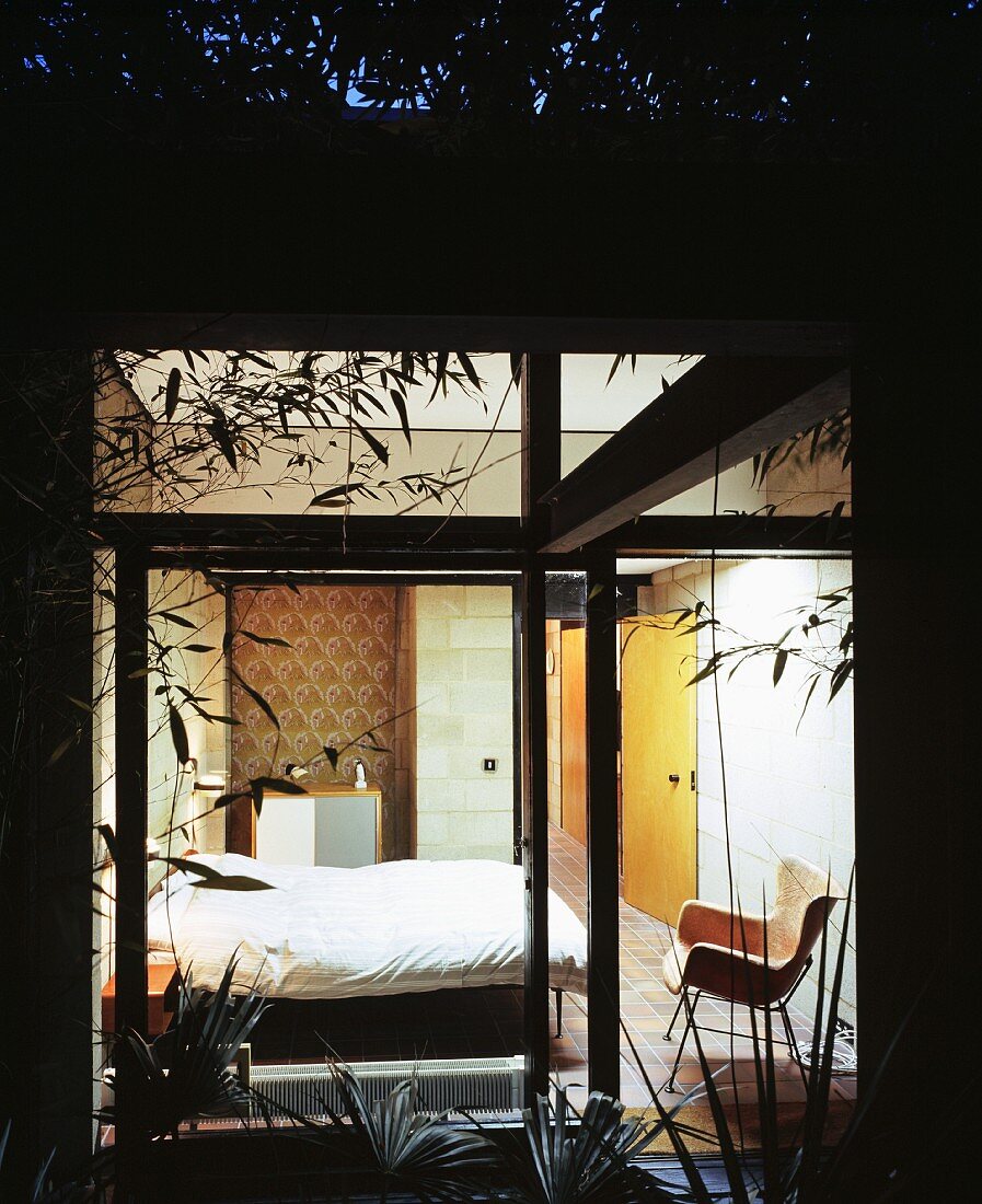 Palms and bamboo leaves in front of an illuminated bedroom window with a retro-style chair