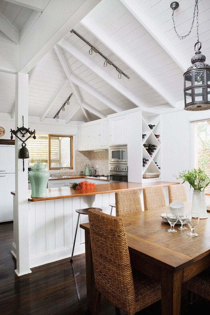 Essplatz mit Holztisch und Rattanstühle vor offener Küche in schlichtem Wohnhaus mit weisser Holzdecke